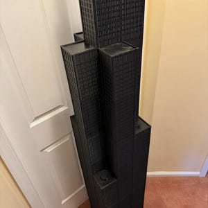 May include: A black architectural model of a skyscraper, featuring multiple rectangular towers of varying heights. The model has a textured surface, with detailed window patterns. The structure is placed on a carpeted floor, near a white door and a beige wall.