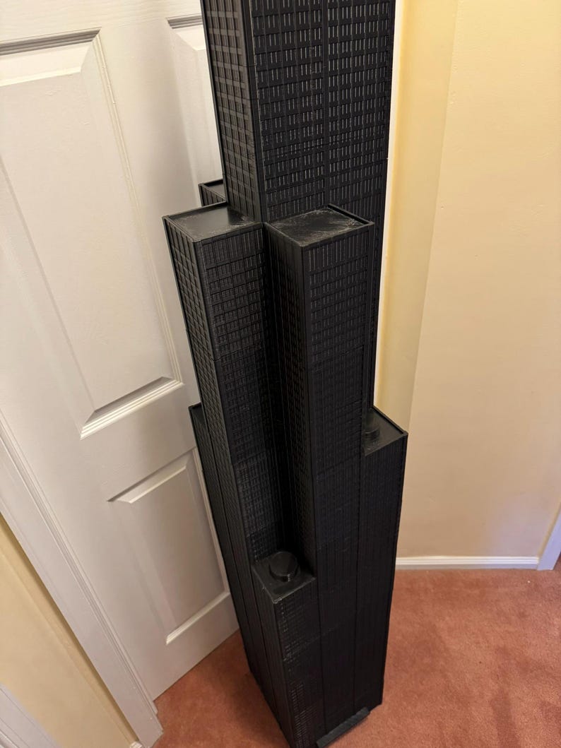 May include: A black architectural model of a skyscraper, featuring multiple rectangular towers of varying heights. The model has a textured surface, with detailed window patterns. The structure is placed on a carpeted floor, near a white door and a beige wall.