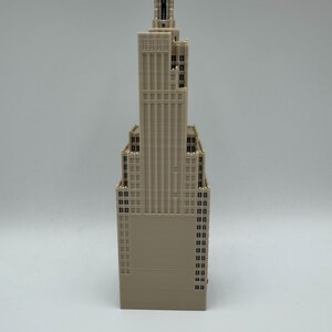 May include: A beige-colored architectural model of a skyscraper. The model features a detailed design with multiple windows and a spire at the top. The building's facade has vertical lines, and the overall structure is tall and slender.
