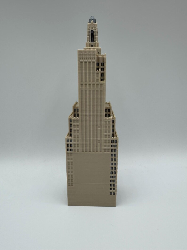 May include: A beige-colored architectural model of a skyscraper. The model features a detailed design with multiple windows and a spire at the top. The building's facade has vertical lines, and the overall structure is tall and slender.
