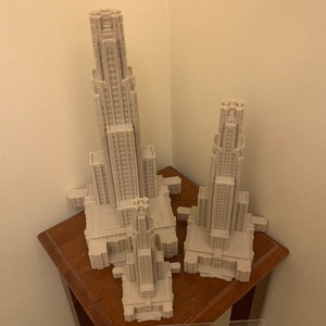 May include: Two 3D printed models of a tall building with a pointed roof and many windows. The models are made of white plastic and are sitting on a wooden table.