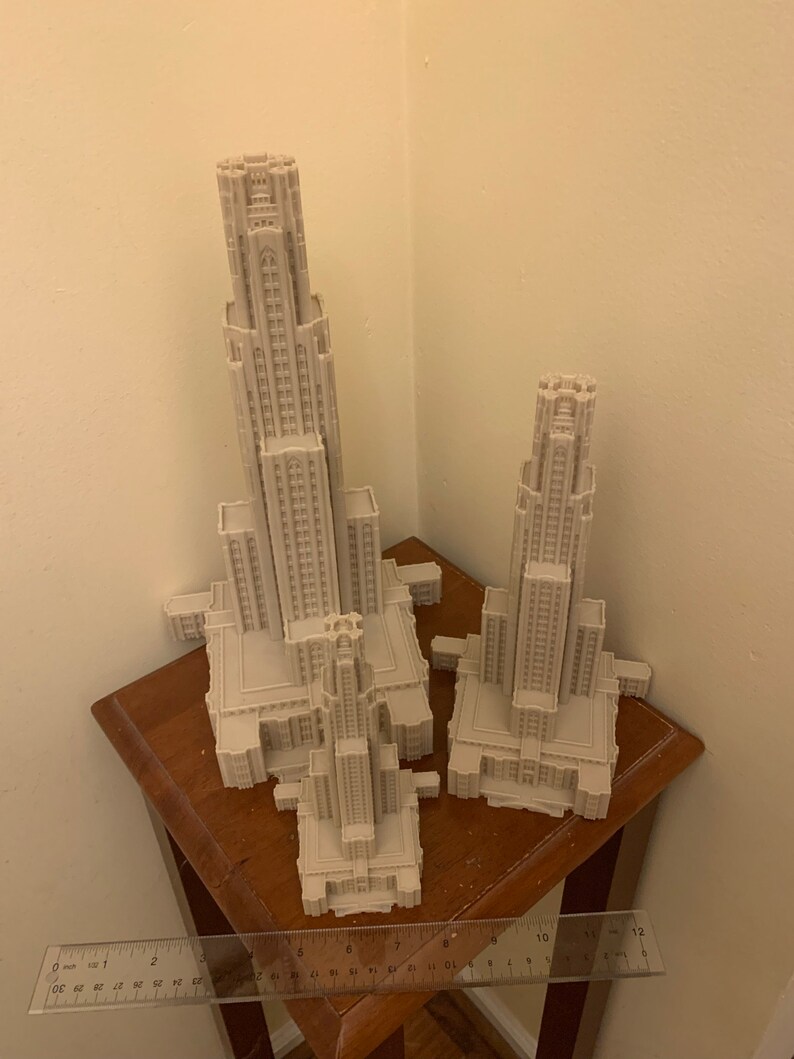 May include: Two 3D printed models of a tall building with a pointed roof and many windows. The models are made of white plastic and are sitting on a wooden table.