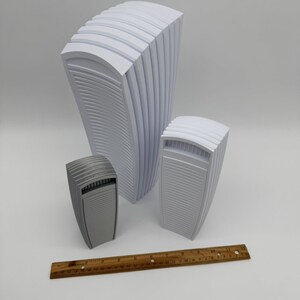 May include: Three abstract, architectural-style sculptures in varying sizes. The largest is white, the medium one is light gray, and the smallest is a darker gray. A wooden ruler with inch markings is at the bottom.