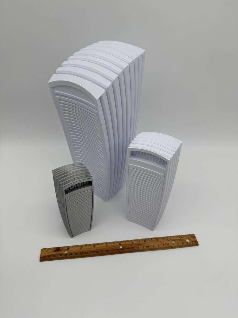 May include: Three abstract, architectural-style sculptures in varying sizes. The largest is white, the medium one is light gray, and the smallest is a darker gray. A wooden ruler with inch markings is at the bottom.
