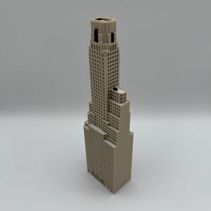 May include: A beige 3D printed model of a skyscraper featuring a stepped architectural design with a tall central tower, multiple rectangular levels, and a grid of small square windows across the exterior surfaces of the building structure.
