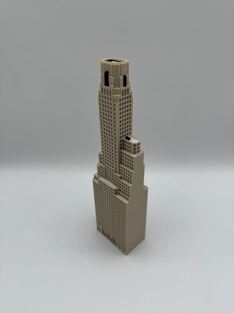 May include: A beige 3D printed model of a skyscraper featuring a stepped architectural design with a tall central tower, multiple rectangular levels, and a grid of small square windows across the exterior surfaces of the building structure.