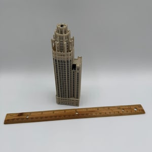 May include: A beige architectural model of a skyscraper with a tiered tower design and numerous vertical window columns. A wooden ruler is placed in front of the model, showing the building stands approximately 7.5 inches in height.