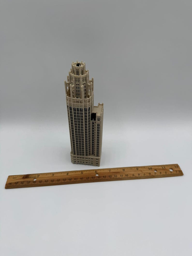 May include: A beige architectural model of a skyscraper with a tiered tower design and numerous vertical window columns. A wooden ruler is placed in front of the model, showing the building stands approximately 7.5 inches in height.