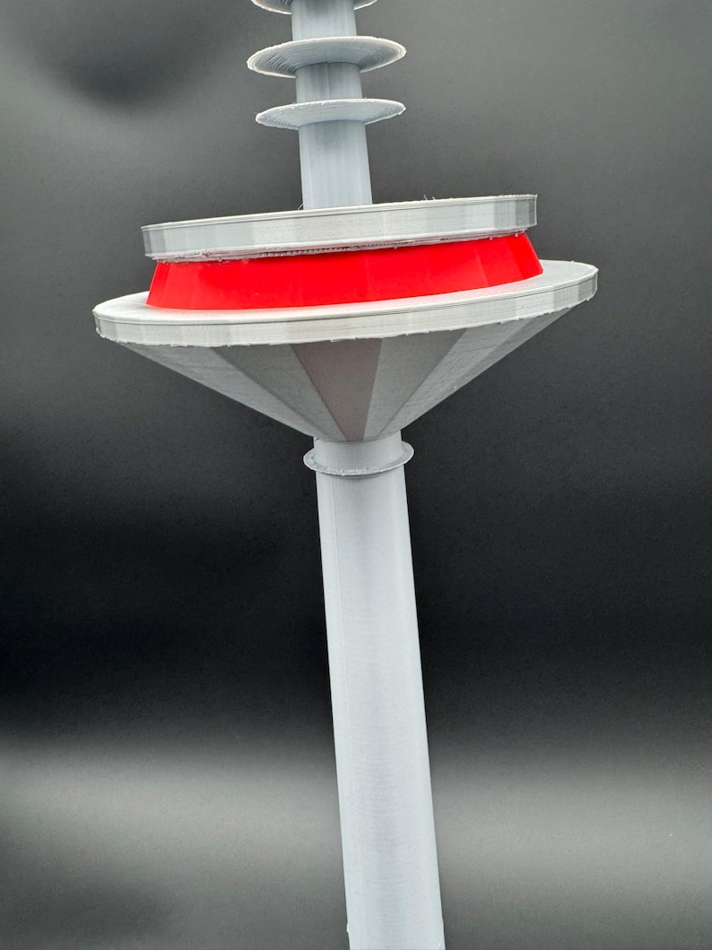 May include: A 3D-printed, abstract sculpture with a light gray stem and a flared, cone-shaped base. A bright red ring sits atop a white disc, with a series of smaller gray discs above. The sculpture is set against a dark background.