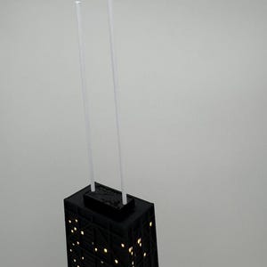 May include: A black architectural model of a skyscraper with illuminated windows. Two white, slender rods extend upwards from the top. The building's facade features a grid-like pattern, and the windows glow with a warm light.