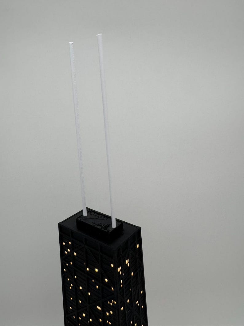 May include: A black architectural model of a skyscraper with illuminated windows. Two white, slender rods extend upwards from the top. The building's facade features a grid-like pattern, and the windows glow with a warm light.