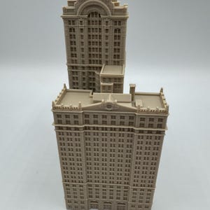 May include: Two beige miniature building models. The taller building has a rectangular shape with many windows and a decorative top. The shorter building is behind the taller one. The models are made of a light-colored material.