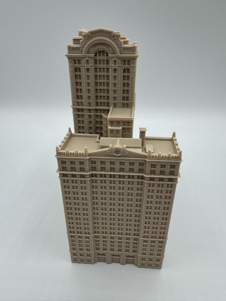 May include: Two beige miniature building models. The taller building has a rectangular shape with many windows and a decorative top. The shorter building is behind the taller one. The models are made of a light-colored material.