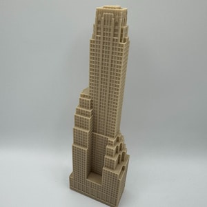 May include: A tan-colored miniature skyscraper model with a detailed facade. The building has a stepped design and a tall, rectangular tower. The model is likely made of a solid material and stands on a white surface.