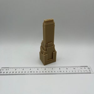 May include: A small, beige, 3D printed model of a skyscraper. The model is about 6 inches tall and is sitting on a white surface. A ruler is in the foreground.