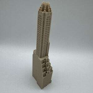 May include: A beige architectural model of a skyscraper. The model features detailed windows and a stepped design on the lower portion. The top of the building has intricate detailing. The model is a miniature representation of a tall building.