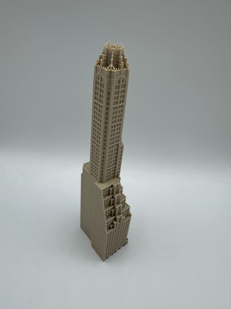 May include: A beige architectural model of a skyscraper. The model features detailed windows and a stepped design on the lower portion. The top of the building has intricate detailing. The model is a miniature representation of a tall building.