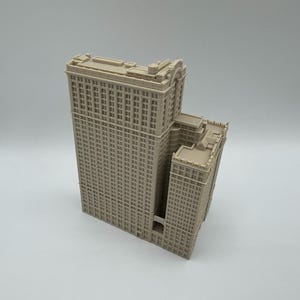 May include: A beige architectural model of a multi-story building with a detailed facade. The model features a tall central tower and a shorter adjacent structure, both with numerous windows and intricate architectural details. The building is set against a plain background.