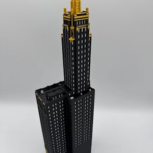 Op de afbeelding: Een zwart en goud model van een wolkenkrabber. Het gebouw heeft een rechthoekige vorm met veel kleine, vierkante ramen. De bovenkant van het gebouw is goudkleurig en het model staat tegen een witte achtergrond.