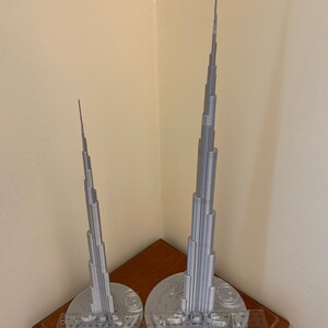 May include: Two silver 3D printed models of the Burj Khalifa, one larger than the other, on a wooden surface with a ruler for scale.