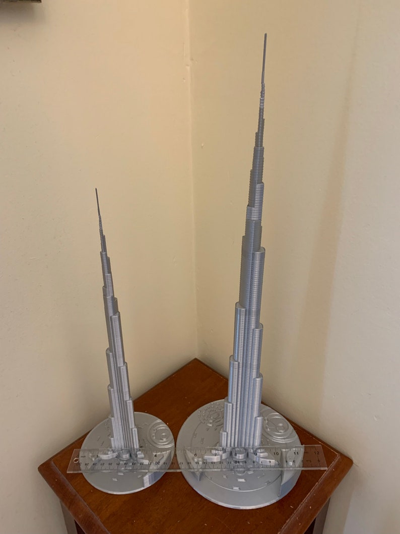 May include: Two silver 3D printed models of the Burj Khalifa, one larger than the other, on a wooden surface with a ruler for scale.