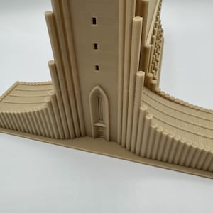 May include: A beige 3D-printed model of a church or cathedral. The model features a tall central tower with rectangular windows, a detailed facade, and a curved base with vertical lines. The architectural model is set against a white background.