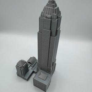 May include: A gray 3D-printed model of a skyscraper with a tall, pointed top and multiple rectangular sections. The model includes adjacent buildings and architectural details, showcasing a modern urban design.
