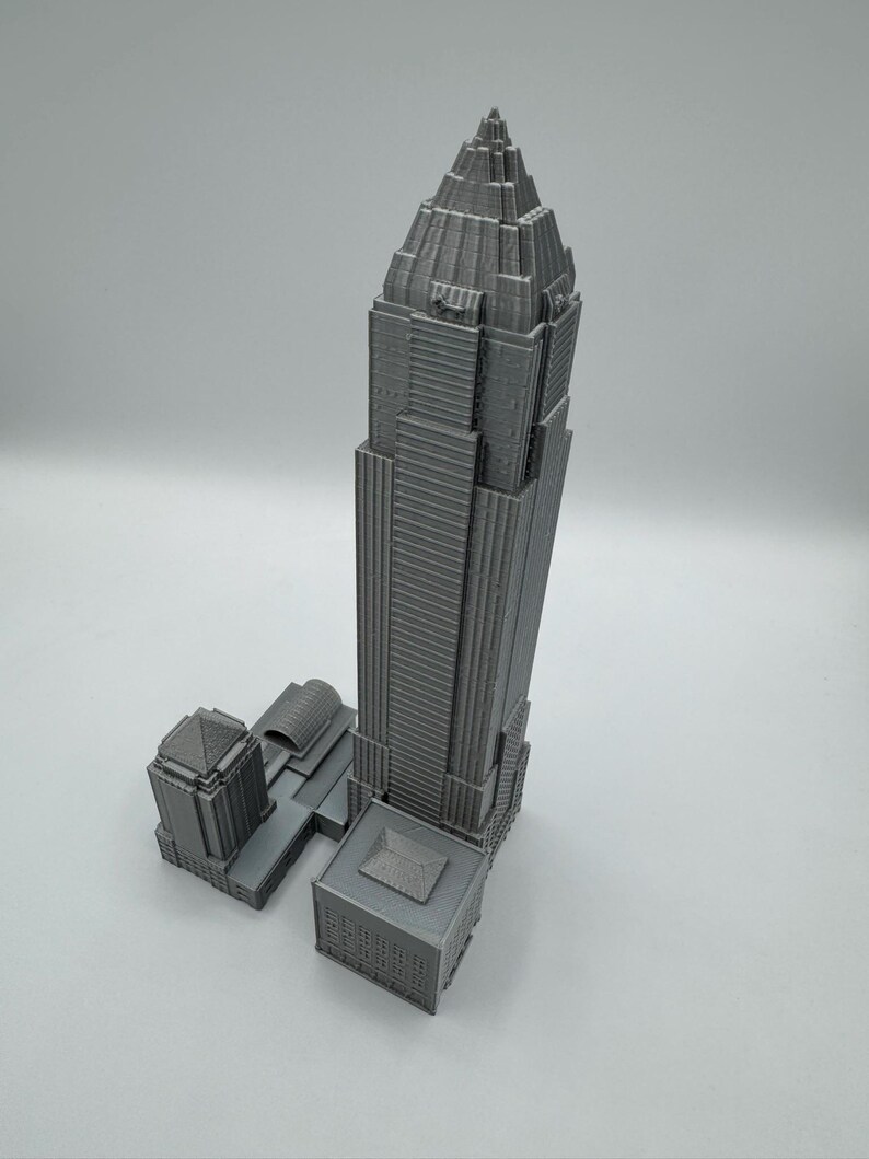 May include: A gray 3D-printed model of a skyscraper with a tall, pointed top and multiple rectangular sections. The model includes adjacent buildings and architectural details, showcasing a modern urban design.