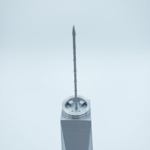May include: A close-up of the top of a gray 3D printed model of a skyscraper. The model has a pointed spire and a square base.