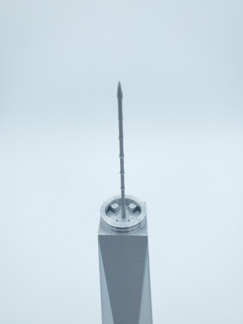 May include: A close-up of the top of a gray 3D printed model of a skyscraper. The model has a pointed spire and a square base.