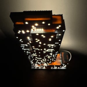 May include: A detailed model of a skyscraper with a lit interior, featuring a brown facade and numerous white lights. The model is made of small, interlocking pieces, creating a realistic representation of a modern building.