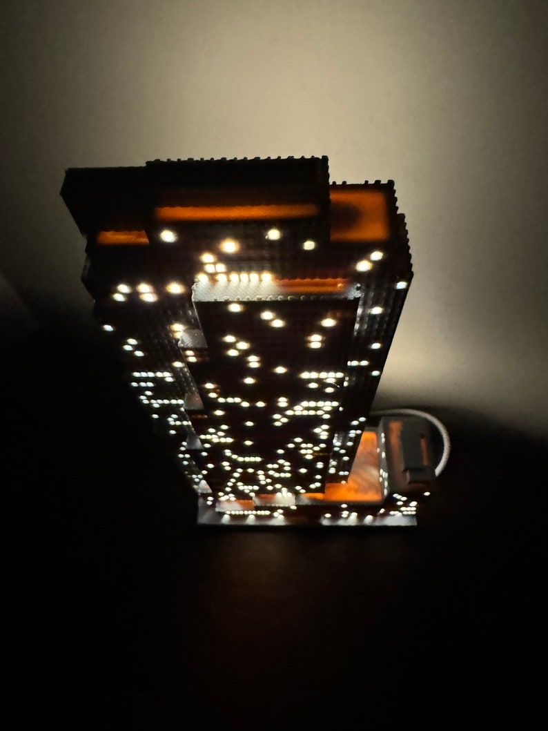 May include: A detailed model of a skyscraper with a lit interior, featuring a brown facade and numerous white lights. The model is made of small, interlocking pieces, creating a realistic representation of a modern building.