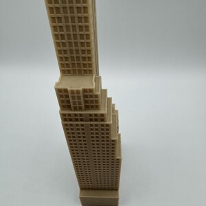 May include: A tan-colored miniature skyscraper model with detailed window patterns. The building has a rectangular base and a tiered design, with a tall, slender tower. The model is likely made of plastic or resin.