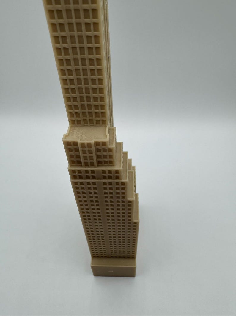 May include: A tan-colored miniature skyscraper model with detailed window patterns. The building has a rectangular base and a tiered design, with a tall, slender tower. The model is likely made of plastic or resin.
