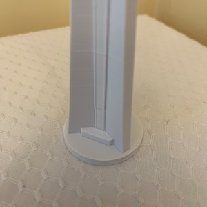 May include: A white, 3D-printed architectural model of a tall, rectangular structure with a circular base. The model has a detailed facade with recessed elements and a small, square platform at the base of the structure.