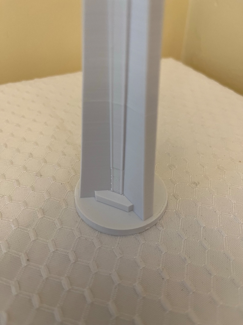 May include: A white, 3D-printed architectural model of a tall, rectangular structure with a circular base. The model has a detailed facade with recessed elements and a small, square platform at the base of the structure.