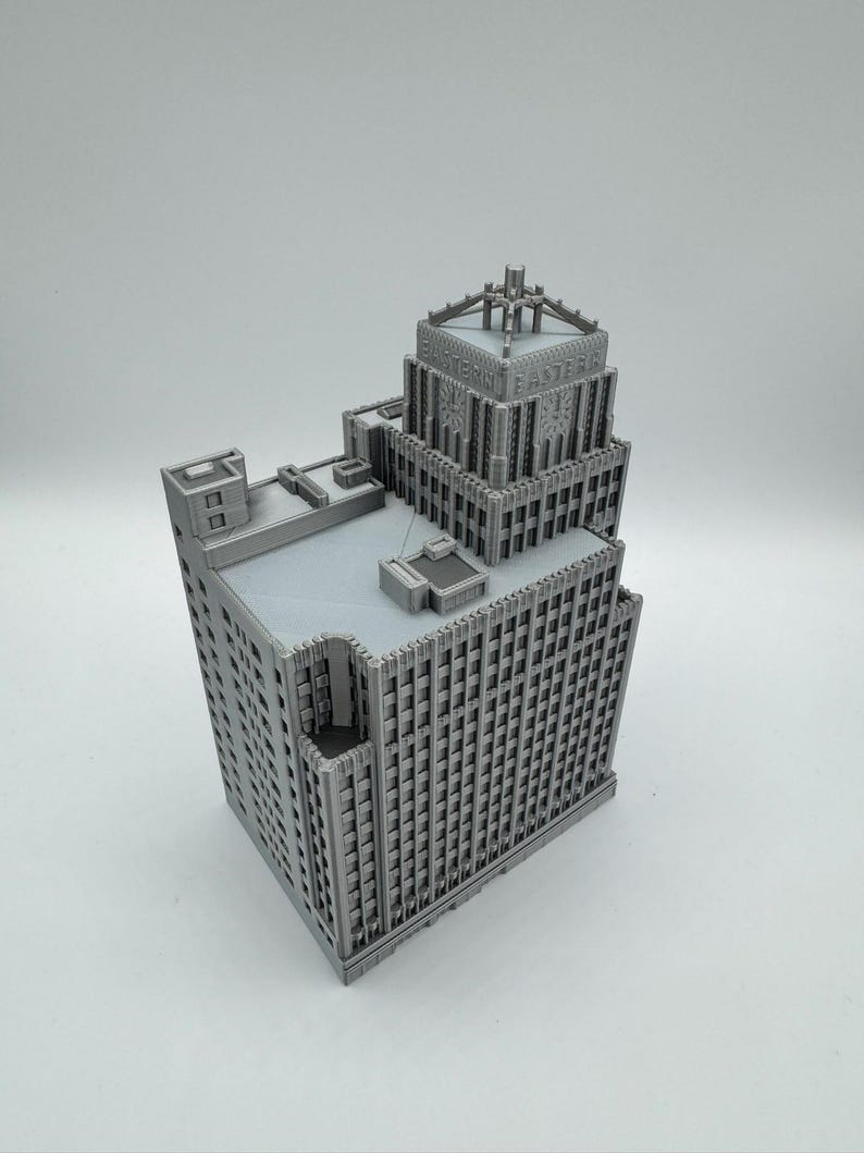 May include: A gray 3D-printed model of a multi-story building with intricate architectural details. The building features a tiered design with numerous windows and a detailed top section. The text "EASTERN" is visible on the top of the building.