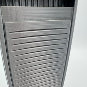 May include: A gray, 3D-printed, rectangular container with a textured surface. The container has horizontal slats on the front and vertical ridges at the top, creating a geometric design. The object is set against a white background.