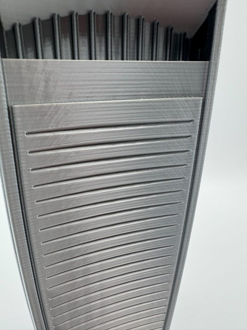 May include: A gray, 3D-printed, rectangular container with a textured surface. The container has horizontal slats on the front and vertical ridges at the top, creating a geometric design. The object is set against a white background.