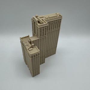 May include: Two beige miniature skyscraper models. The larger building has a rectangular shape with many windows and a detailed top. The smaller building is similar in design, with a flat roof and a rectangular shape.