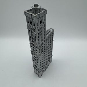 May include: A grey miniature model of a tall building, featuring intricate architectural details and multiple storeys. The building has a rectangular main structure with an attached smaller section. The model is likely made of plastic or similar material.