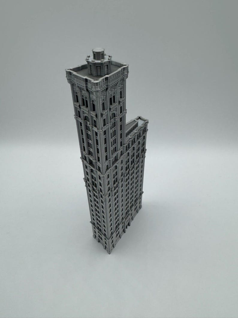 May include: A grey miniature model of a tall building, featuring intricate architectural details and multiple storeys. The building has a rectangular main structure with an attached smaller section. The model is likely made of plastic or similar material.