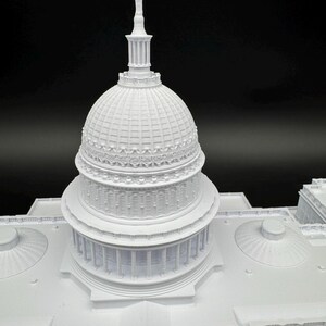 K&ouml;nnte beinhalten: Ein detailliertes, wei&szlig;es 3D-gedrucktes Modell eines Geb&auml;udes mit einer gro&szlig;en Kuppel und S&auml;ulen. Das Modell weist komplizierte architektonische Details auf, darunter eine hohe Spitze auf der Kuppel. Das Geb&auml;ude steht vor einem schwarzen Hintergrund.