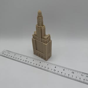 May include: A beige miniature model of a tall building with a domed roof. The model is approximately 7 inches tall and is sitting on a white surface next to a ruler.