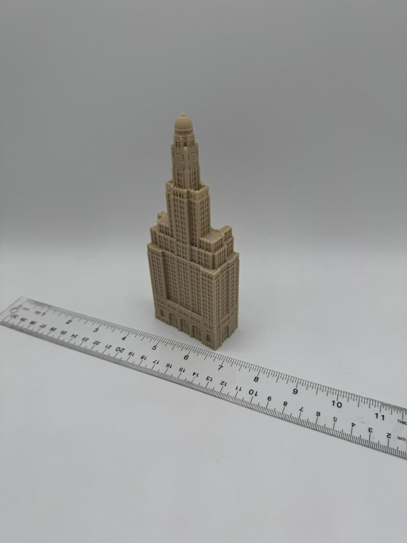 May include: A beige miniature model of a tall building with a domed roof. The model is approximately 7 inches tall and is sitting on a white surface next to a ruler.