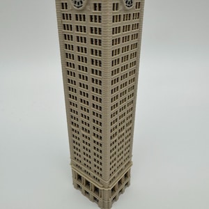 May include: A beige 3D printed model of a tall skyscraper with a rectangular base and numerous windows. The structure features a prominent clock face near the top on two visible sides, with black clock hands and numerals set against a light background.