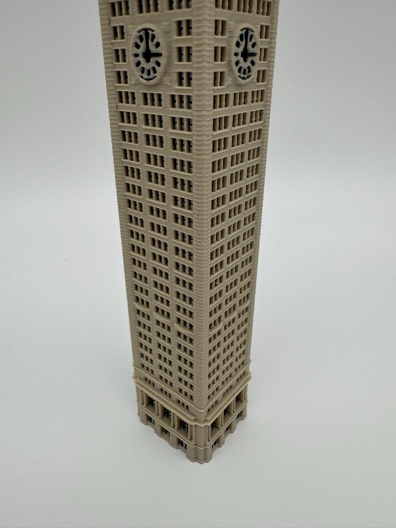 May include: A beige 3D printed model of a tall skyscraper with a rectangular base and numerous windows. The structure features a prominent clock face near the top on two visible sides, with black clock hands and numerals set against a light background.