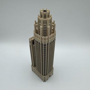 May include: A beige 3D printed architectural model of a skyscraper featuring a tiered, neo-Gothic design with intricate vertical columns, recessed windows, and a central tower structure that tapers toward a crenelated top with a dark opening.
