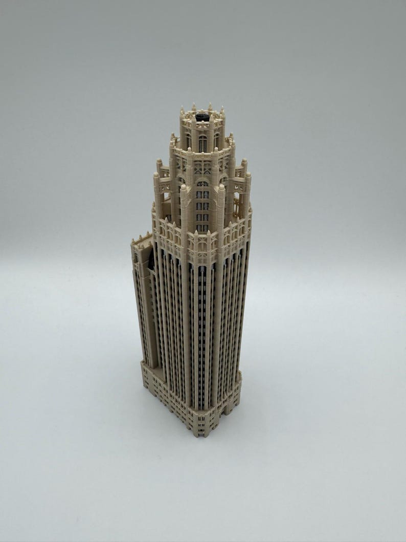 May include: A beige 3D printed architectural model of a skyscraper featuring a tiered, neo-Gothic design with intricate vertical columns, recessed windows, and a central tower structure that tapers toward a crenelated top with a dark opening.