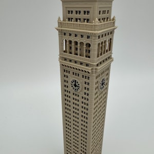 May include: A beige, three-dimensional printed model of a tall clock tower building. The structure features numerous rectangular windows, a prominent clock face on two visible sides, and an ornate, multi-tiered top section with arched openings.